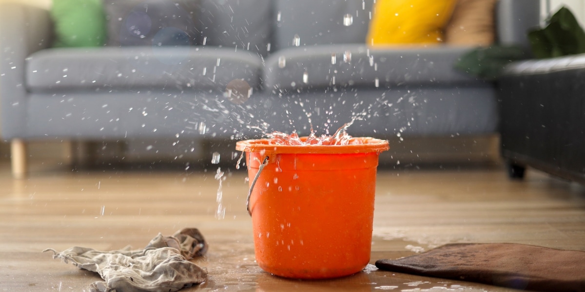 An orange bucket catches water dripping from a leaking ceiling, with towels and splashes on the floor. Highlights the need for emergency roof repair services provided by Priority Roofing.